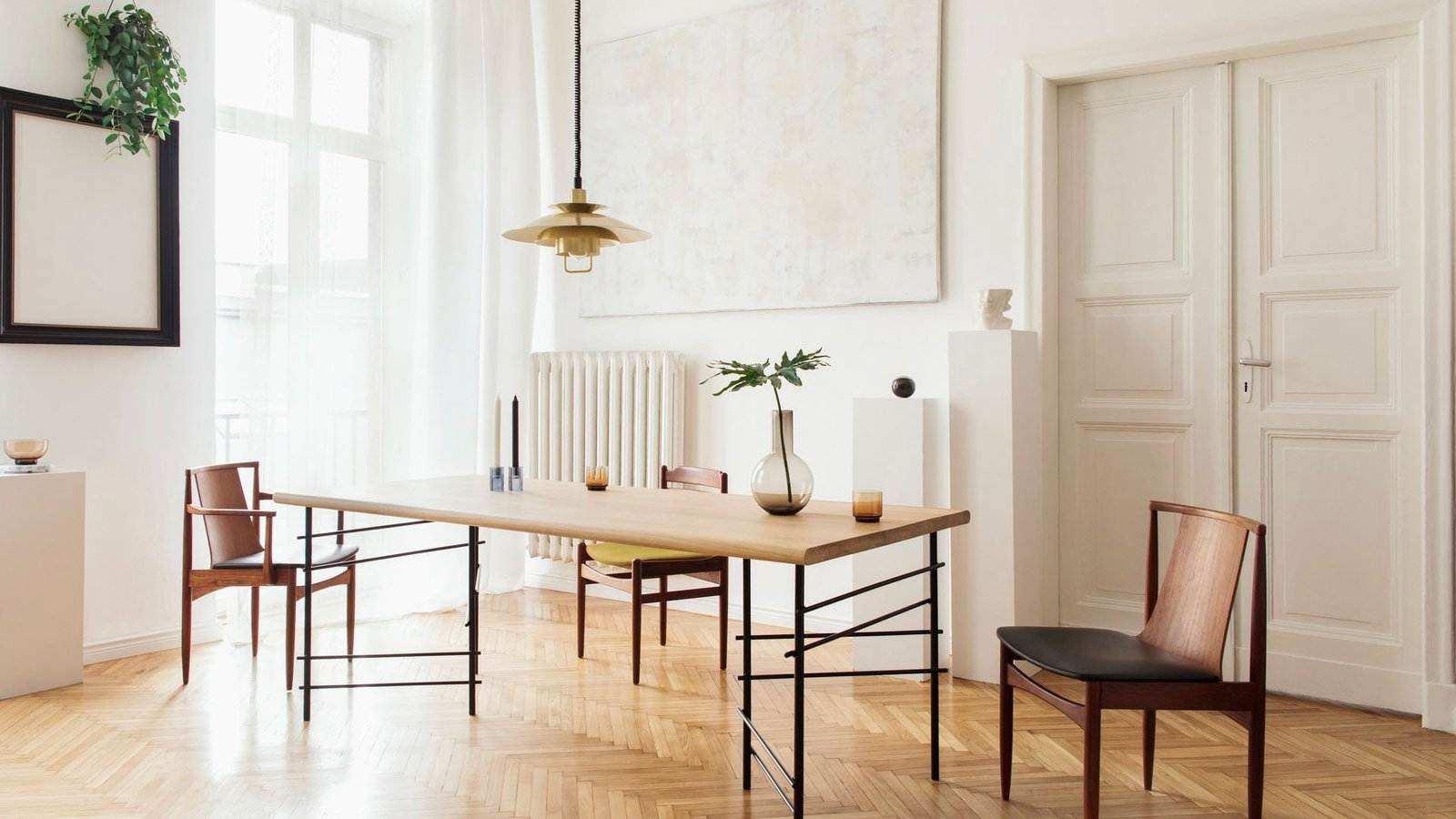 Ein minimalistisches Esszimmer mit einem rechteckigen Holztisch und vier Holzstühlen. Über dem Tisch hängt eine Pendelleuchte. Der Raum verfügt über ein großes Fenster mit transparenten Vorhängen, einen hellen Fischgrätboden, abstrakte Kunst an einer weißen Wand und Dekoartikel auf Podesten.