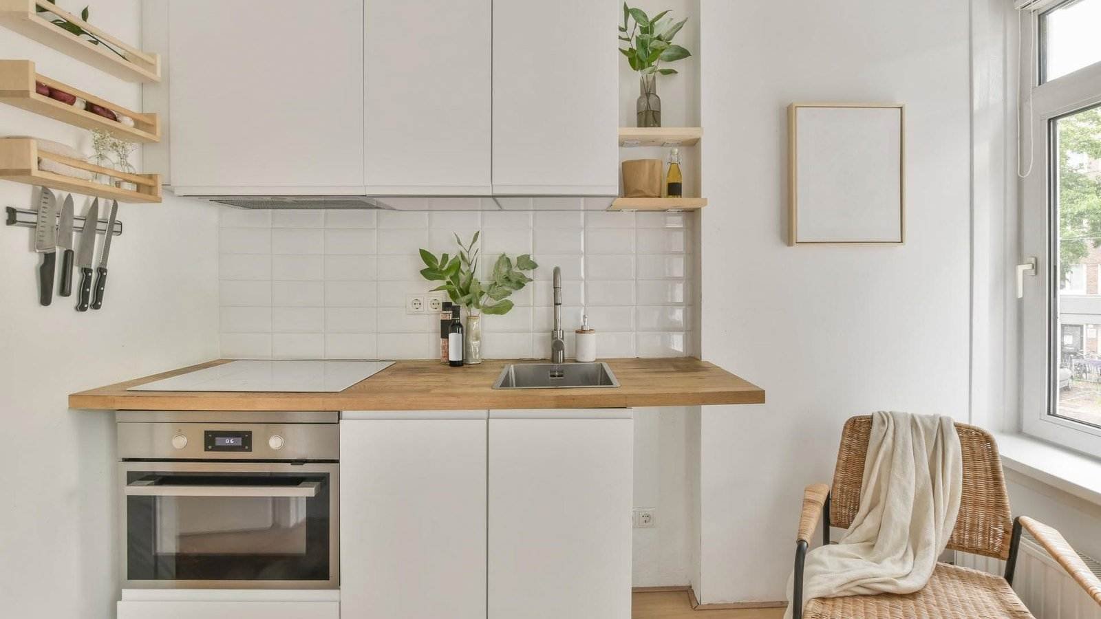 Moderne Küche mit weißen Schränken und Holzarbeitsplatte. Spüle, Elektroherd und Backofen sind integriert. Messer hängen an der Wand. Regale bieten Platz für Pflanzen und Gefäße. Ein Korbstuhl mit einer drapierten Decke steht am Fenster und sorgt für Tageslicht. Die Wände sind weiß gefliest.