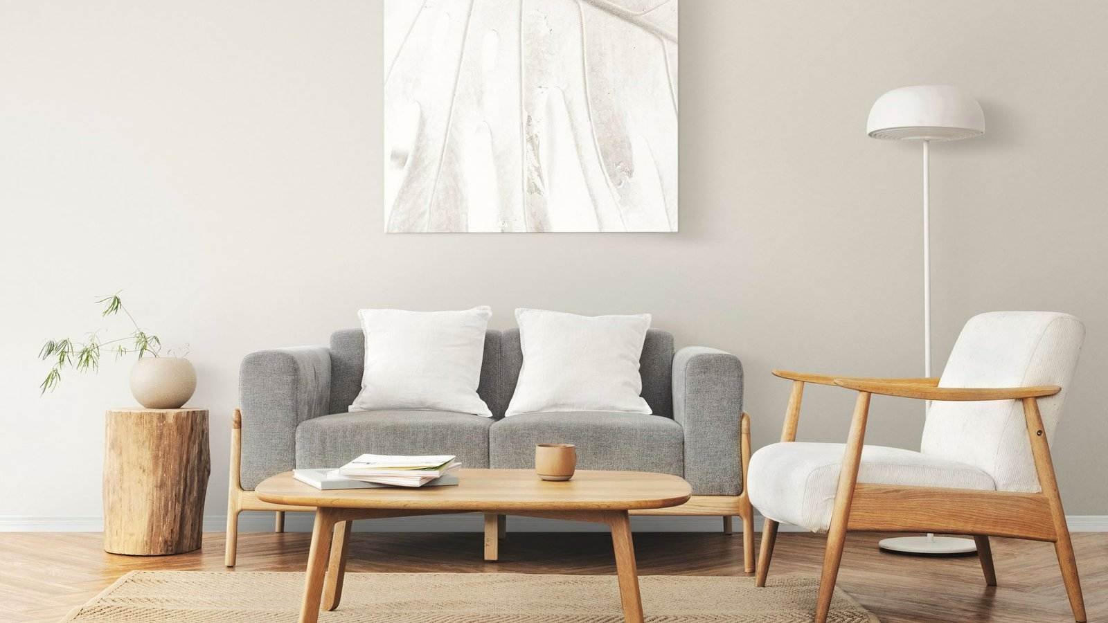 Ein minimalistisches Wohnzimmer verfügt über ein graues Sofa mit zwei weißen Kissen, einen hellen Holzsessel mit weißem Kissen und einen ovalen Couchtisch aus hellem Holz. Auf einem kleinen runden Beistelltisch aus Holz steht eine Vase mit Grünpflanzen. Eine hohe Stehlampe und abstrakte Wandkunst runden das Dekor ab.